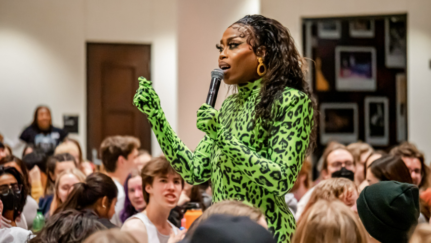 Drag queen walking with microphone speaking to students in the Great Hall