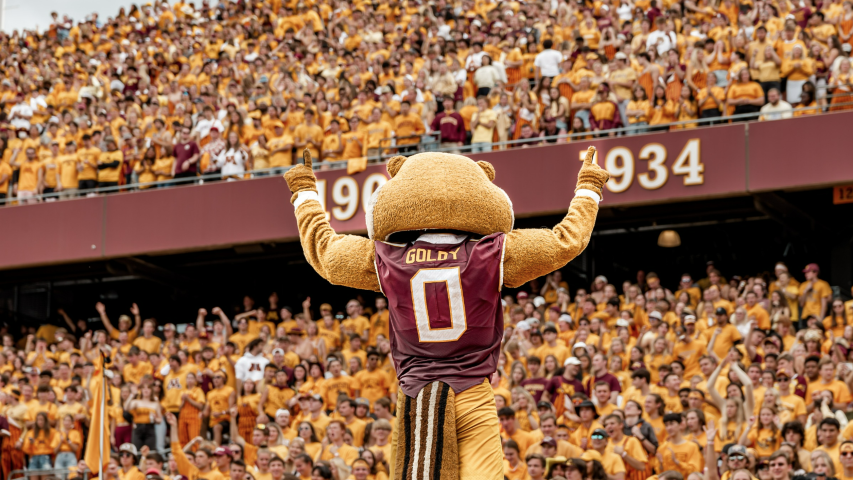 Goldy the Gopher mascot raising hands in front of the Homecoming crowd in football stadium